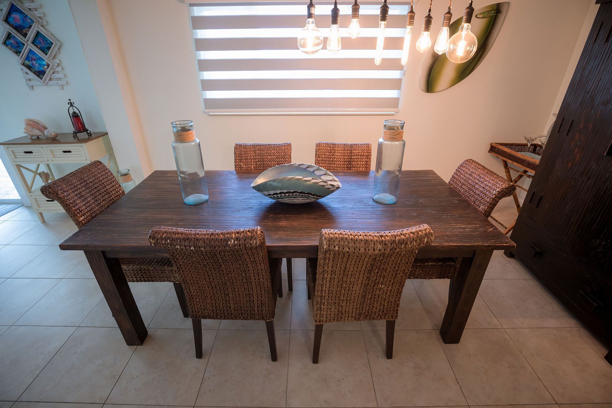 A dining area with a wooden table and wicker chairs, a decorative bowl, and a wall-mounted light fixture.