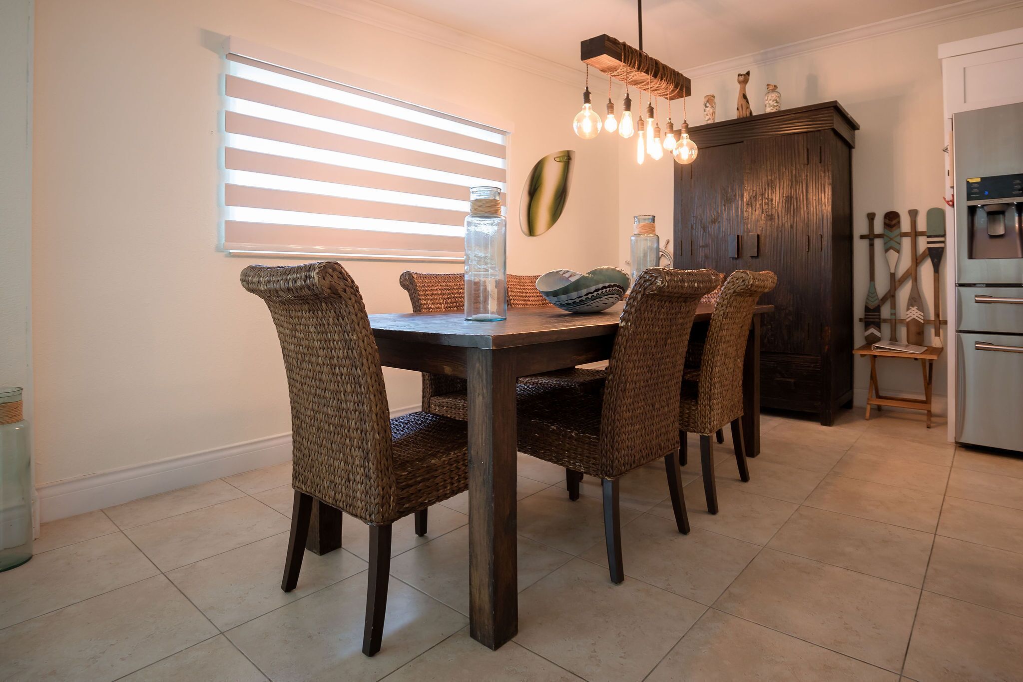 A dining area with a dark wooden table and wicker chairs, a hanging light fixture, and a window with blinds.