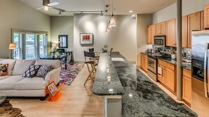 A modern kitchen with granite countertops, wooden cabinets, and stainless steel appliances. There is a dining area with a large table and chairs, and a living room with a sofa and a coffee table.