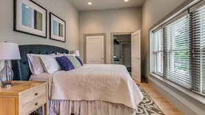 A bedroom with a bed, bedside table, lamp, and a patterned rug. There are two framed pictures on the wall, a closet, and a window with blinds.