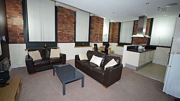 A living room with a brown leather sofa, a glass coffee table, and a kitchen area with white cabinets and a brick wall. - Premier Apartments (Bradford)