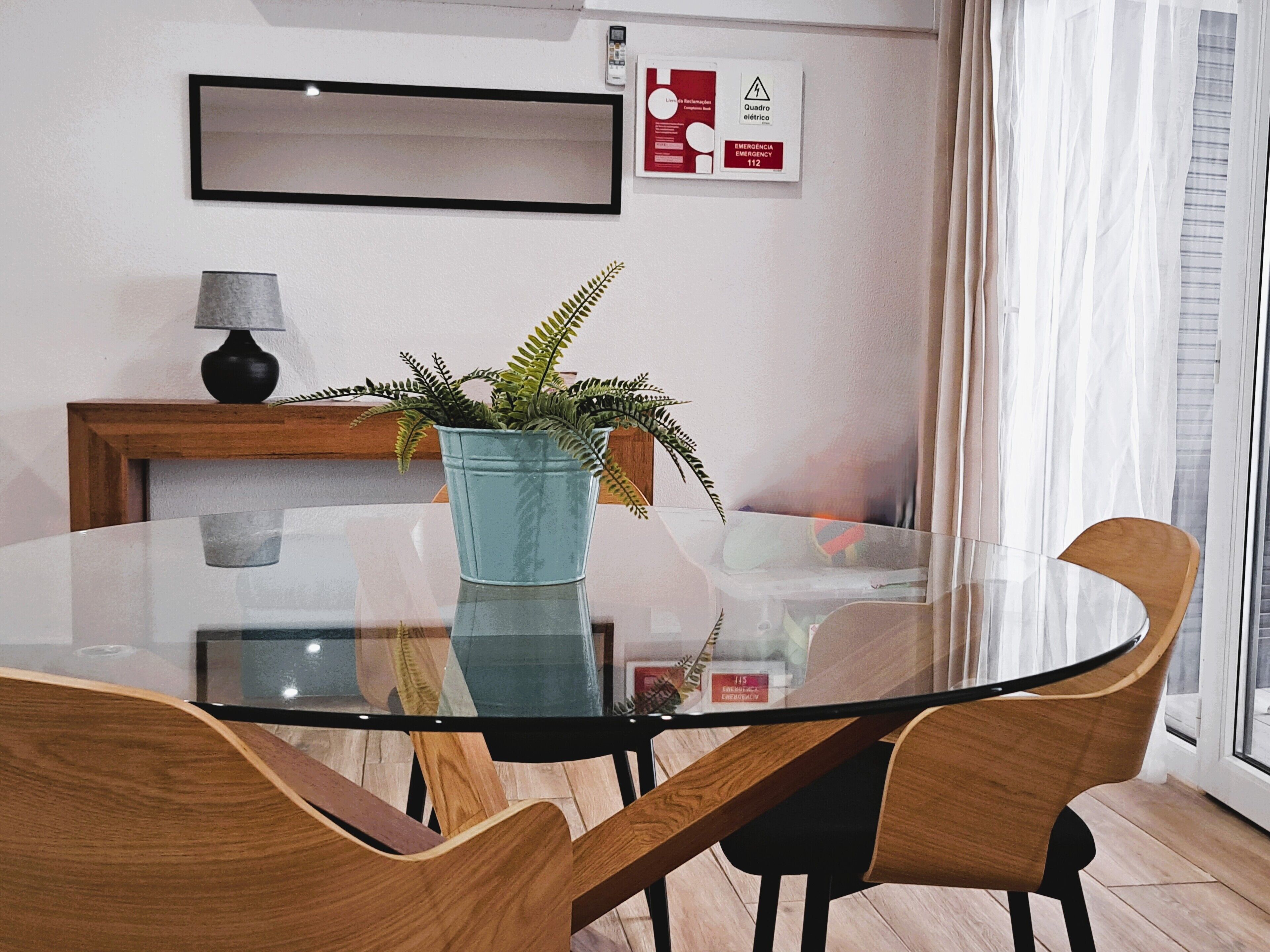 Een moderne kamer met een glazen tafel, omringd door houten stoelen, een potplant met een varen en een lamp op een houten bijzettafeltje.