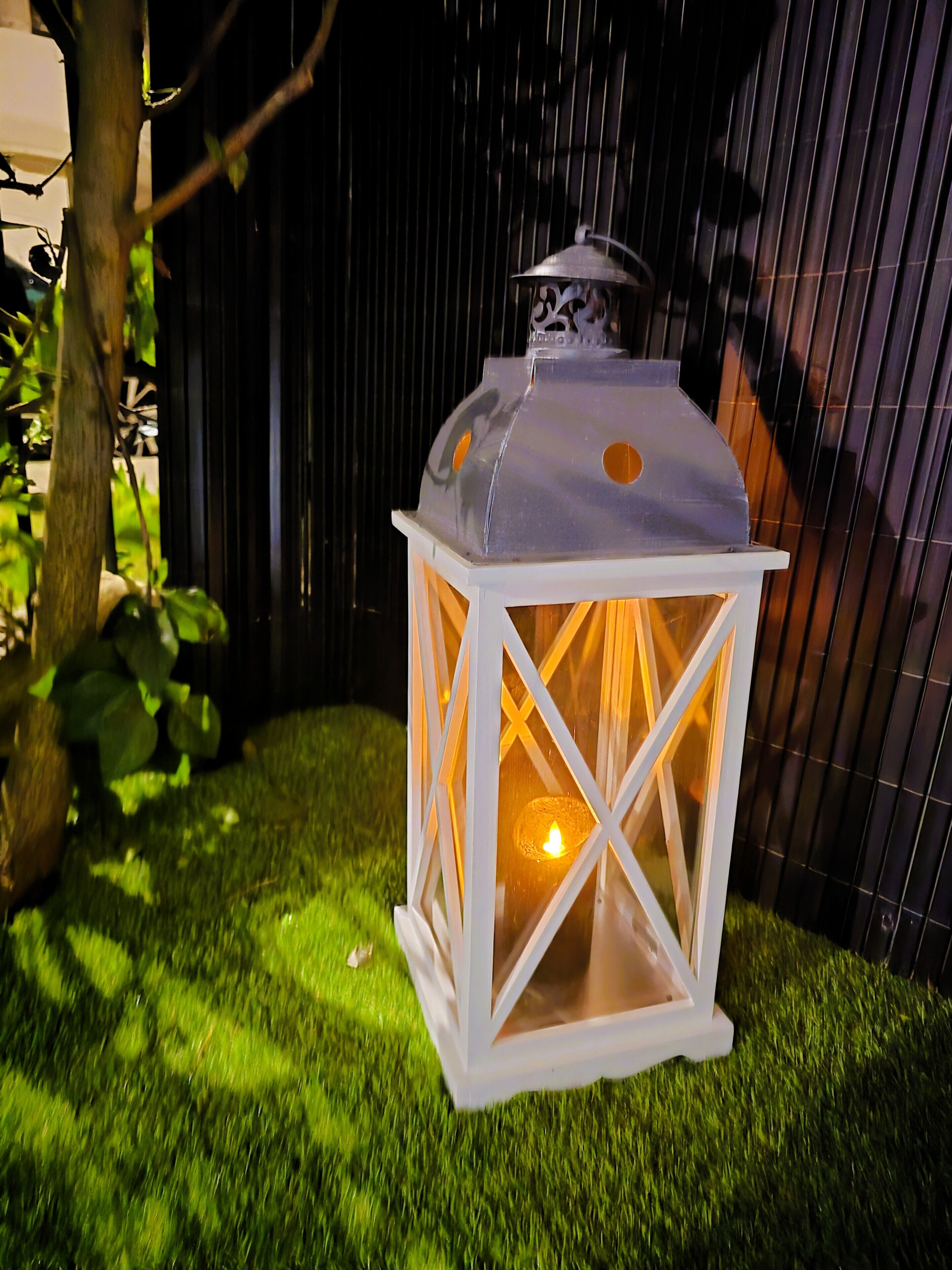A white lantern with a lit candle inside, placed on artificial grass.