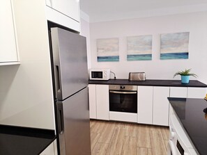 Cozinha moderna com eletrodomésticos de aço inoxidável, armários brancos, bancada preta e vista para o oceano através de uma obra de arte.