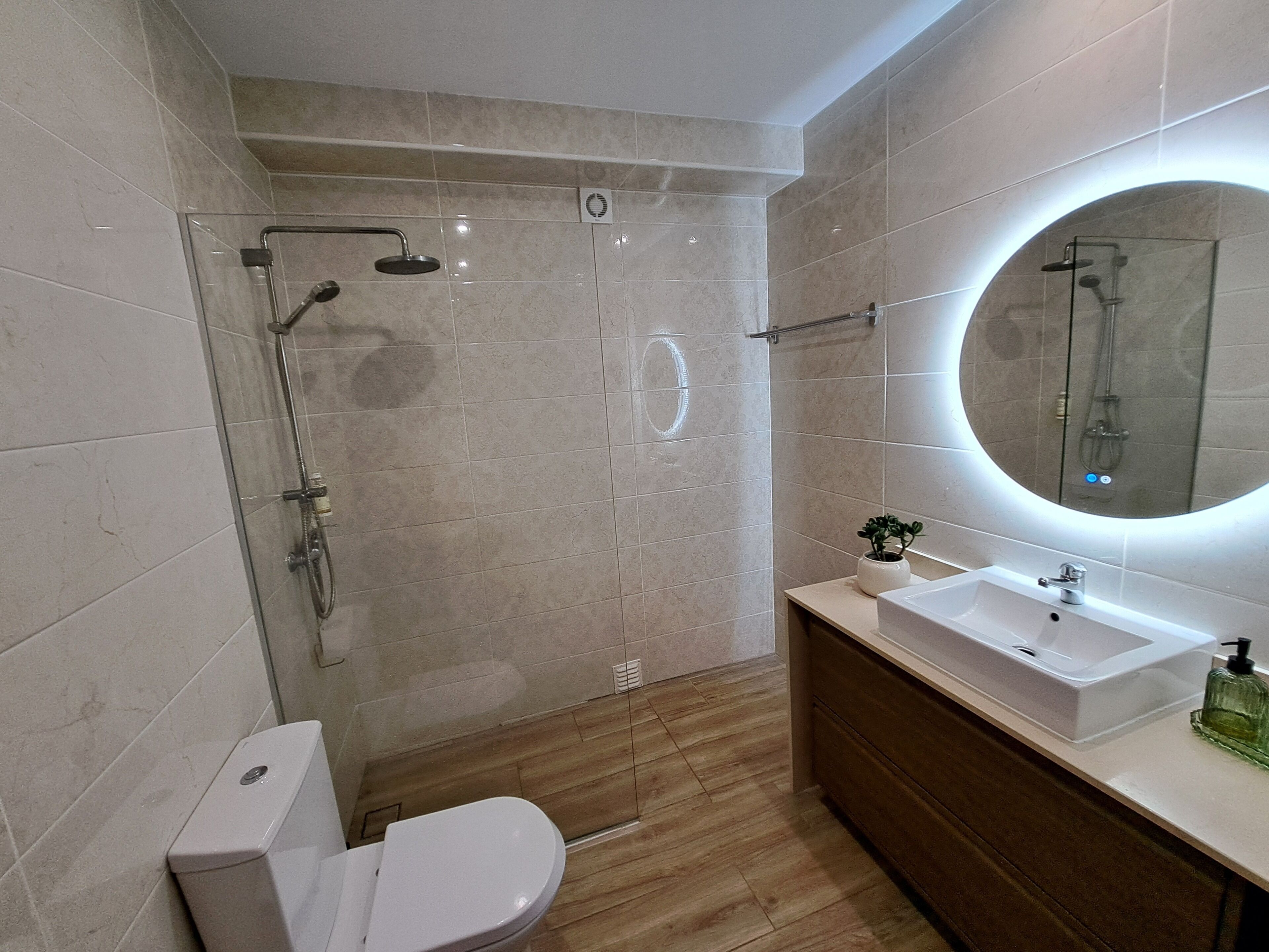 A modern bathroom with a glass-enclosed shower, a white toilet, a wooden vanity with a sink, and a mirror with a circular light.