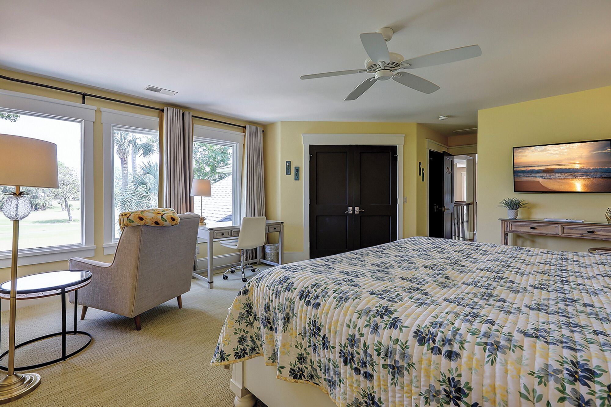 Une chambre d’hôtel équipée d’un lit, d’un bureau, d’une chaise, d’un ventilateur de plafond et d’une grande fenêtre avec des rideaux.