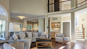 A spacious living room with a high ceiling, a staircase leading to an upper level, and a kitchen area visible in the background.