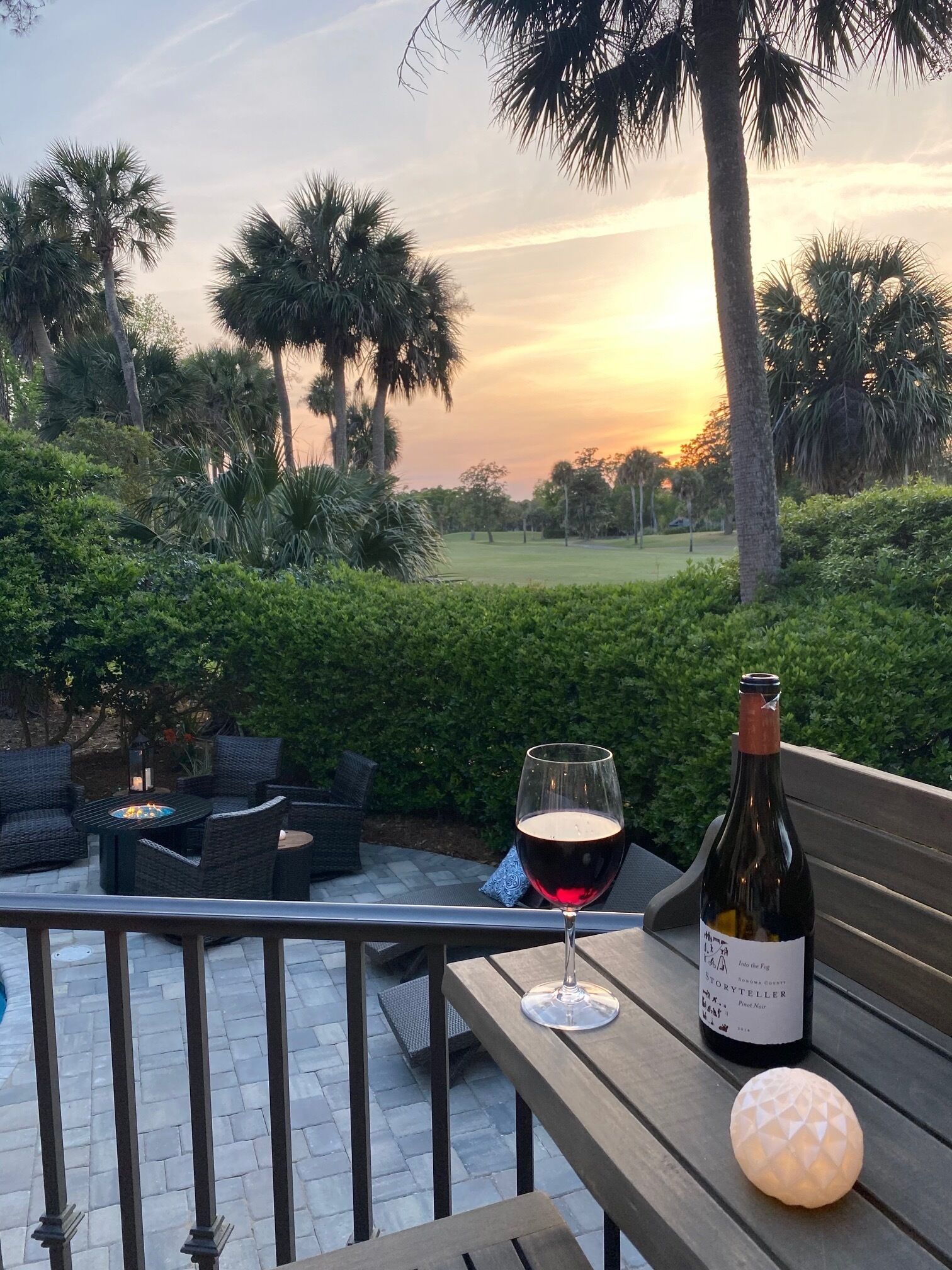 An outdoor patio with wicker furniture, a glass of red wine, a bottle of wine, and a decorative light on a table.