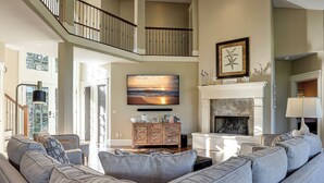 A spacious living room with a high ceiling, a fireplace, and a large flat-screen TV mounted on the wall.