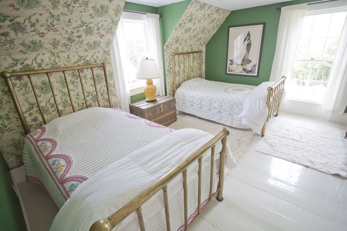 A bedroom with two beds, floral wallpaper, a painting, and a window with curtains.