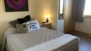 Un lit bien fait, avec un oreiller sur lequel est brodé le message « Faites quelque chose de bien aujourd’hui », une lampe de chevet, un miroir et un grand tableau représentant une fleur violette accroché au mur.