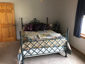 A bedroom with a metal bed frame, a wooden door, a window with curtains, and a bedside table with a lamp and flowers.