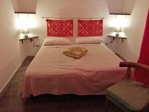 A single bed with a pink bedspread, two white pillows, and a beige throw pillow. There are two wall-mounted lamps on either side of the bed. A wooden chair with a green cushion is positioned to the right of the bed.