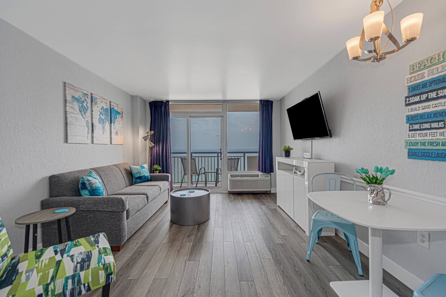 A modern living room with a grey sofa, a small round table, a TV mounted on the wall, and a balcony with a view.