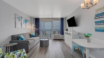A modern living room with a grey sofa, a small round table, a TV mounted on the wall, and a balcony with a view.