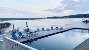Un área de piscina con sillones de descanso y una mesa, con vistas a una masa de agua tranquila con un muelle y barcos al fondo.