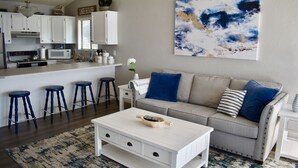 Cocina moderna con gabinetes blancos, un microondas y un lavamanos. Hay una barra con taburetes y una mesa de centro blanca con un bol encima. Un sofá gris con almohadas azules está en la sala de estar, y un gran cuadro abstracto adorna la pared.