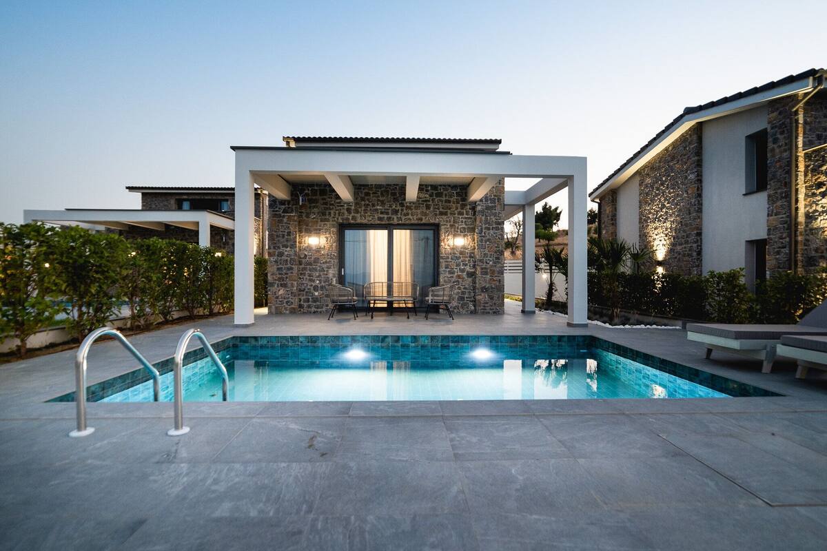 A modern outdoor pool area with a stone wall, two lounge chairs, and a pergola.