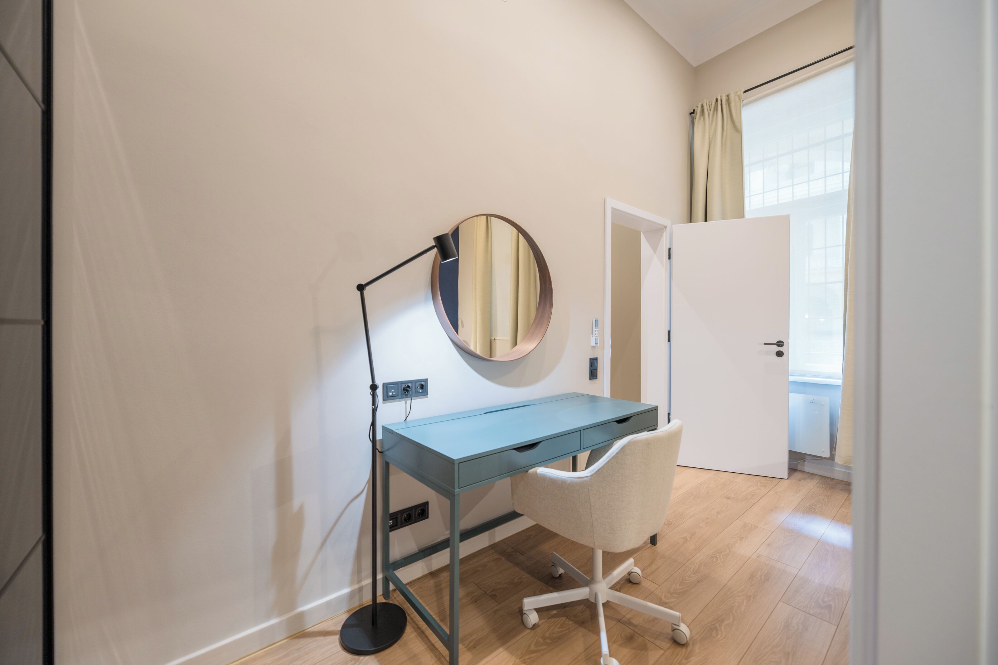 A modern room with a glass desk, a round mirror, a white chair, and a door leading to a balcony.