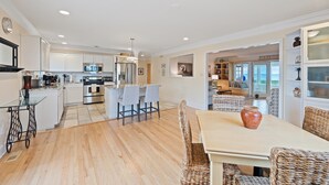 A modern kitchen with white cabinetry, stainless steel appliances, and a dining area with wicker chairs and a table. - Beachfront Home-Fully Furnished.  PRIVATE Beach Rights on L.I. Sound. (Mt Sinai)