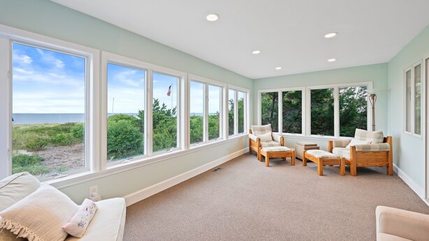 A spacious room with large windows offering a view of the beach and greenery. There are multiple seating areas with light-colored furniture. - Beachfront Home-Fully Furnished.  PRIVATE Beach Rights on L.I. Sound. (Mt Sinai)