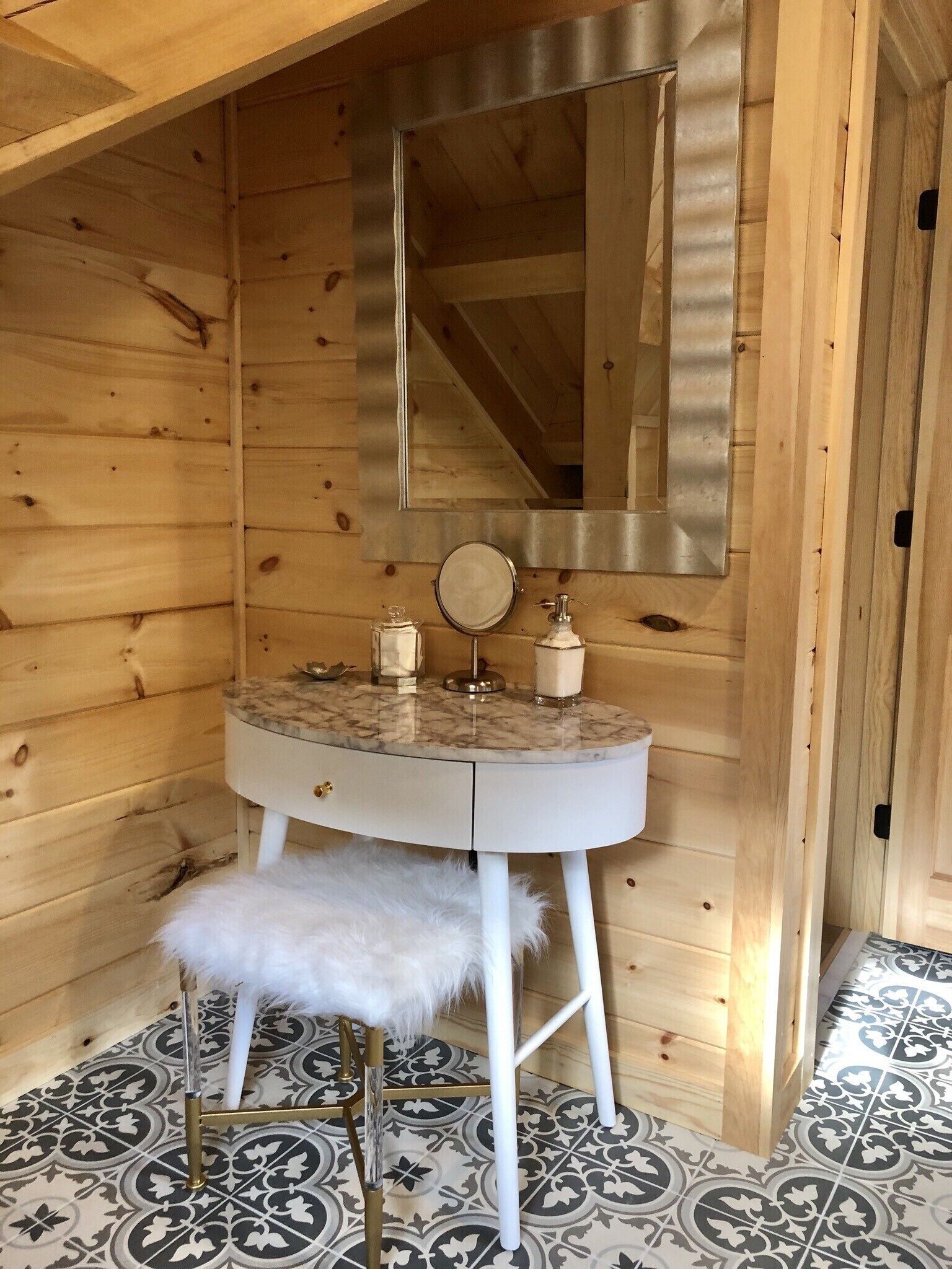 Ein Holzblockhaus-Interieur mit einem Marmorwaschtisch, einem Spiegel, einem kleinen runden Tisch, einem weißen, flauschigen Hocker und gemusterten Bodenfliesen.