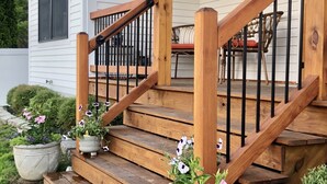 Eine Holztreppe führt zu einer Veranda mit Geländer, Blumentöpfen und einem Essbereich mit Korbstühlen.