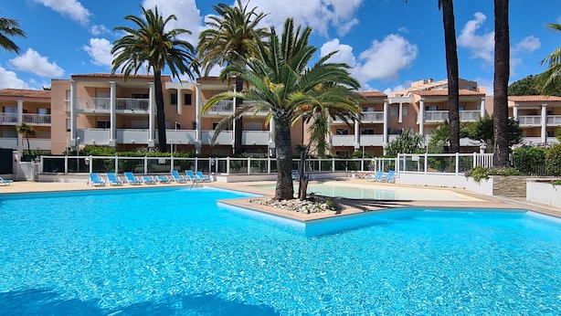 Une piscine d’un bleu éclatant, bordée de chaises longues et de palmiers, le tout entouré d’un immeuble à plusieurs étages doté de balcons.