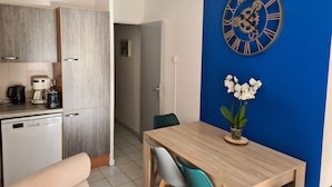 Une cuisine moderne équipée d’un lave-vaisselle, d’une machine à café et d’une bouilloire. On y trouve un coin repas avec une table et des chaises en bois, ainsi qu’une horloge murale avec des chiffres romains.