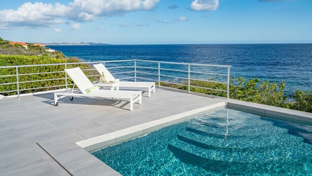 A pool with a view of the ocean, white lounge chairs, and a railing. - Villa 308 - Coral Estate (Rif. St. Marie)