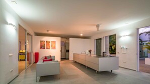 A modern kitchen with a central island, a dining area with a white sofa, and a view of the outdoors through a sliding door. - Villa 308 - Coral Estate (Rif. St. Marie)