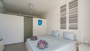 A bedroom with a bed, bedside tables, a ceiling fan, a window with blinds, and a door leading to another room. - Villa 308 - Coral Estate (Rif. St. Marie)