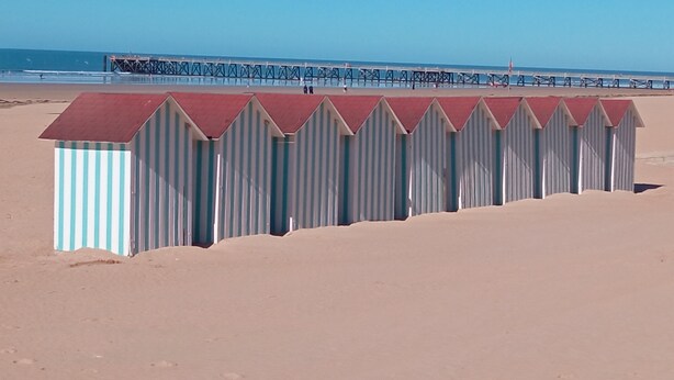 A row of beach huts with a metal roof and striped walls, located on a sandy beach with a pier in the background. - HOUSE WITH HEATED POOL CLOSE TO BEACHES AND NATIONAL FOREST (Saint-Jean-de-Monts, Vendée)