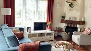 A living room with a blue sofa, white armchair, and a TV on a white cabinet. There are wicker lamps, a fireplace, and a window with curtains. - DETACHED HOUSE IN THE CENTER OF VALE DAS FURNAS  (Furnas)