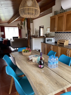 Un coin repas avec une table en bois, des chaises bleues, un luminaire suspendu en fibres tressées, et une cuisine en arrière-plan.