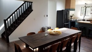 Un comedor con mesa y sillas de madera, una escalera, una cocina con refrigerador y estufa, y un ventanal con luz natural.