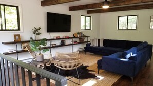 Una sala de estar con un sofá azul, una silla de mimbre, un televisor montado en la pared y una estantería con libros y objetos decorativos.