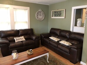 A living room with two brown leather sofas, a wooden coffee table, a wall clock, and a 'Home sweet home' sign.