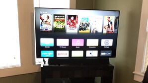A living room with a flat-screen TV on a stand, a brown leather couch, a wooden coffee table, and a glass candle holder. - Historic Downtown, Wifi, Apple TVs, Cold AC (Brownsville)