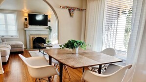 Uma sala de estar moderna com uma mesa de jantar de madeira, cadeiras brancas, uma TV de tela plana, lareira e uma janela grande com persianas.