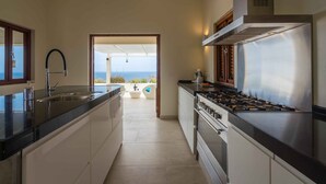A modern kitchen with a large island, stainless steel appliances, and a view of the sea through an open doorway. - Villa Vila Blenchi - Coral Estate (Rif. St. Marie)