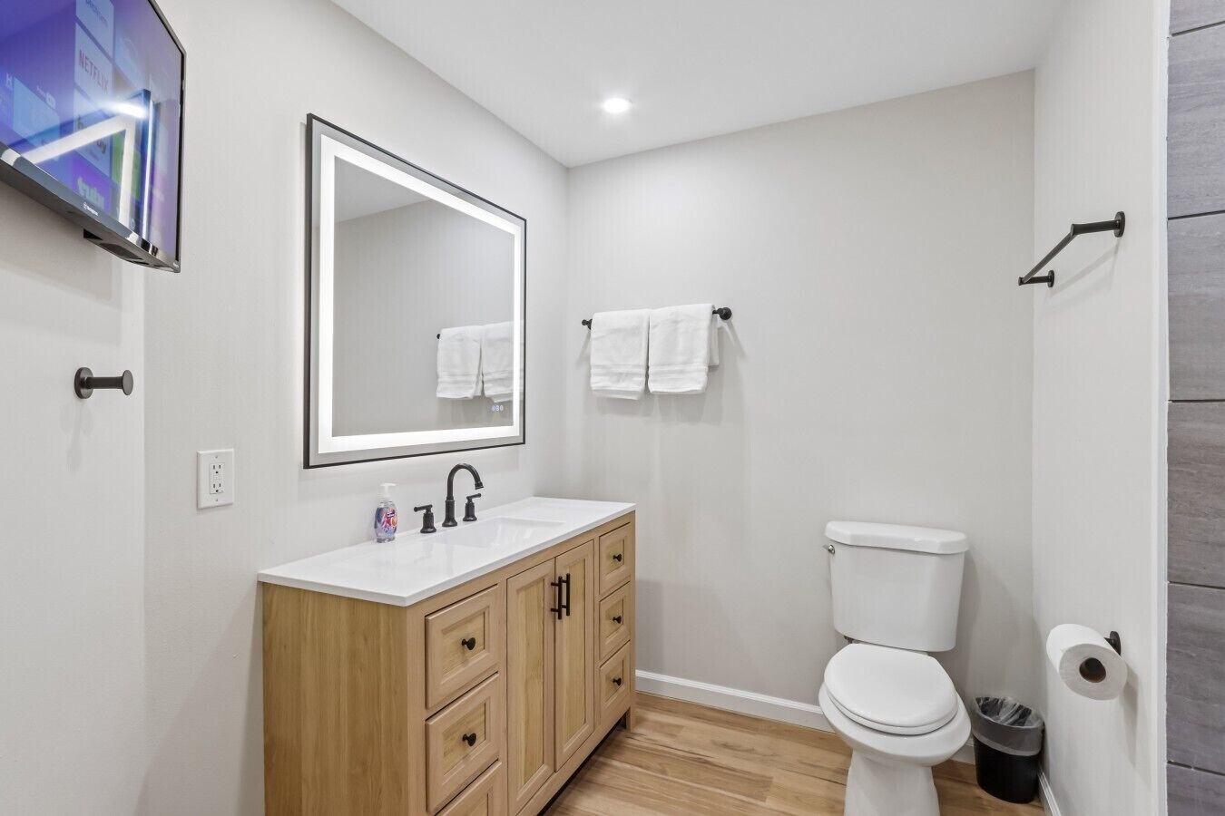 A modern bathroom with a white sink, wooden vanity, a large mirror, a toilet, and a wall-mounted TV.