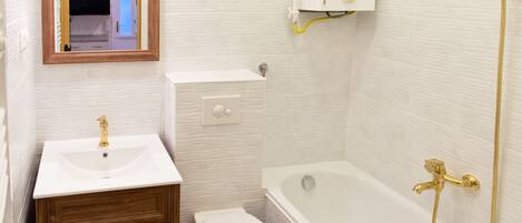 Un baño con lavamanos blanco, tocador de madera, inodoro blanco y bañera con cabezal de ducha dorado.