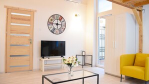 A living room with a wooden door, a clock, a flat-screen TV, a small table with a vase, a yellow armchair, and a window with a view. - Cosy flat with balcony (Budapest)
