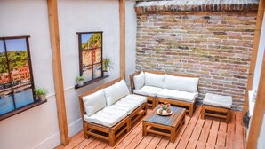 A wooden deck with a sofa set and a small table, a brick wall, and a window with a cityscape view. - Cosy flat with balcony (Budapest)