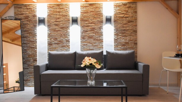 A modern living room with a stone wall, a grey sofa, a glass coffee table, and a vase with flowers. - Cosy flat with balcony (Budapest)