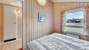 A bedroom with wooden panel walls, a bed with white bedding, a framed picture on the wall, and a window with a view of a house and trees. - Amazing home in Hvide Sande with sauna (Hvide Sande)