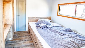 Una stanza piccola con un letto singolo, pavimento in legno, una porta azzurra e una finestra con cornice in legno.