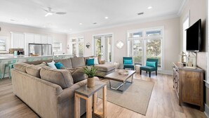 A spacious living room with a large sofa, a coffee table, and two armchairs. There is a kitchen area with white cabinets and a refrigerator. The room has large windows and a ceiling fan. - Gulf View - 30A Private Heated Pool, 3 min walk to the beach. No roads to cross. (Santa Rosa Beach)
