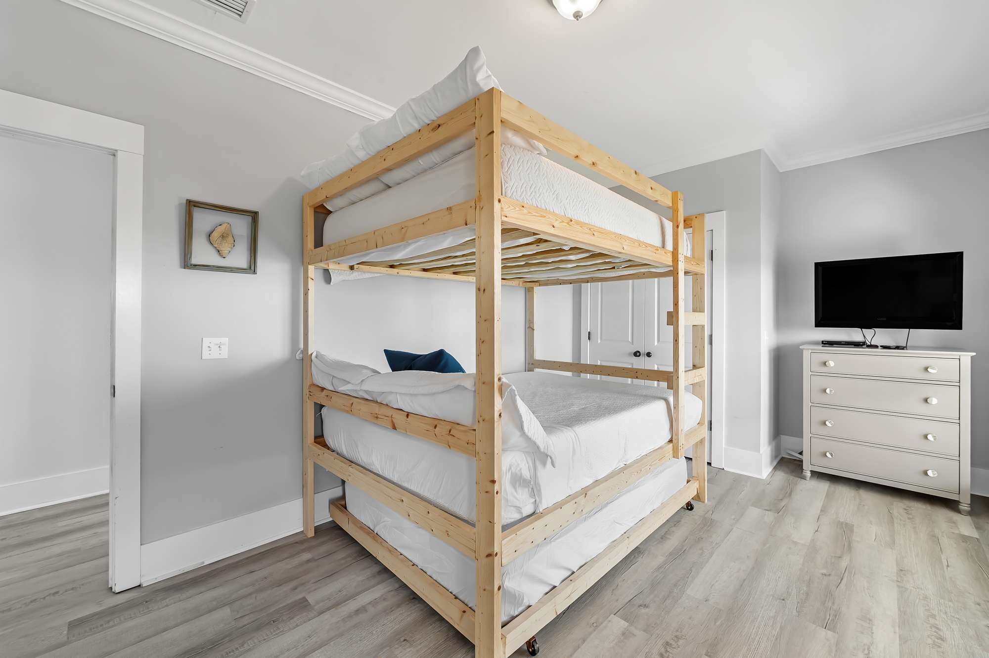 A bunk bed with white bedding, a dresser with drawers, a television, and a framed picture on the wall.